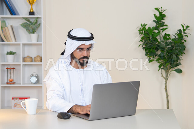 إدارة شؤون الأعمال وتنظيمها، احتساء مشروب القهوة، استخدام جهاز تقني حديث ومتطور، رجل عربي خليجي اماراتي يرتدي الكندورة والغترة البيضاء يستخدم الحاسوب، مهنة ووظيفة مكتبية، دمج العمل مع التقنية والتكنولوجيا، تطور الشركات الاماراتية، إتمام مهام العمل في المكتب