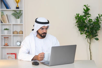 تطور الشركات الاماراتية، إتمام مهام العمل في المكتب، إدارة شؤون الأعمال وتنظيمها، احتساء مشروب القهوة، استخدام جهاز تقني حديث ومتطور، رجل عربي خليجي اماراتي يرتدي الكندورة والغترة البيضاء يستخدم الحاسوب، مهنة ووظيفة مكتبية، دمج العمل مع التقنية والتكنولوجيا