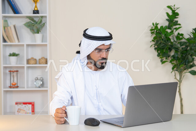 A Saudi woman using a computer in the office, using a modern and advanced technical device, drinking her favorite drink, gestures of concentration and integration, managing business and completing required tasks, a female profession and job, a veiled Arab Gulf Emirati woman wearing a colorful abaya sitting behind a table and drinking coffee