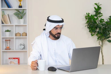 A Saudi woman using a computer in the office, using a modern and advanced technical device, drinking her favorite drink, gestures of concentration and integration, managing business and completing required tasks, a female profession and job, a veiled Arab Gulf Emirati woman wearing a colorful abaya sitting behind a table and drinking coffee