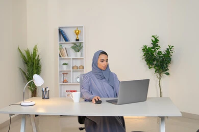 ايماءات التركيز والاندماج، ادارة الأعمال وانجاز المهام المطلوبة، مهنة ووظيفة نسائية،  إمرأة عربية خليجية اماراتية محجبة ترتدي عباءة ملونة تجلس خلف الطاولة، سيدة سعودية تستخدم الحاسوب في المكتب، استخدام جهاز تقني حديث ومتطور