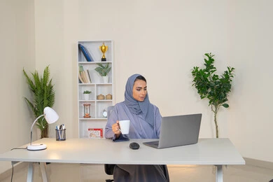 A female profession and job, a veiled Emirati Gulf Arab woman wearing a colorful abaya sitting behind a table, a Saudi woman using a computer in the office, using a modern and advanced technical device, gestures of concentration and integration, business management and completing required tasks