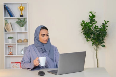 ايماءات التركيز والاندماج، ادارة الأعمال وانجاز المهام المطلوبة، مهنة ووظيفة نسائية،  إمرأة عربية خليجية اماراتية محجبة ترتدي عباءة ملونة تجلس خلف الطاولة، سيدة سعودية تستخدم الحاسوب في المكتب، استخدام جهاز تقني حديث ومتطور