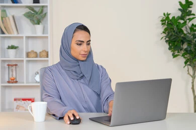 مهنة ووظيفة نسائية،  إمرأة عربية خليجية اماراتية محجبة ترتدي عباءة ملونة تجلس خلف الطاولة، سيدة سعودية تستخدم الحاسوب في المكتب، استخدام جهاز تقني حديث ومتطور، ايماءات التركيز والاندماج، ادارة الأعمال وانجاز المهام المطلوبة
