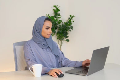 ادارة الأعمال وانجاز المهام المطلوبة، مهنة ووظيفة نسائية،  إمرأة عربية خليجية اماراتية محجبة ترتدي عباءة ملونة تجلس خلف الطاولة، سيدة سعودية تستخدم الحاسوب في المكتب، استخدام جهاز تقني حديث ومتطور، ايماءات التركيز والاندماج
