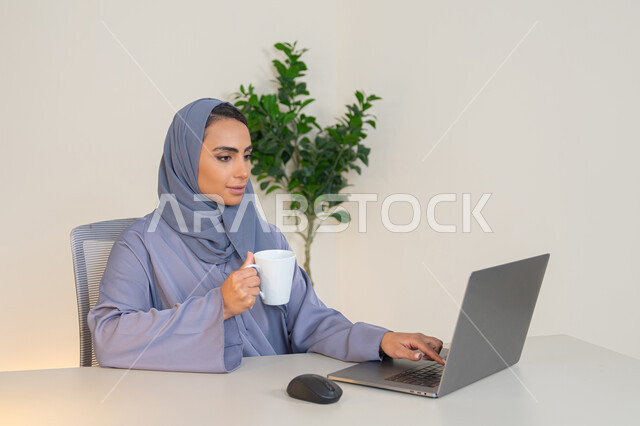 سيدة سعودية تستخدم الحاسوب في المكتب، استخدام جهاز تقني حديث ومتطور، احتساء المشروب المفضل، ايماءات التركيز والاندماج، ادارة الأعمال وانجاز المهام المطلوبة، مهنة ووظيفة نسائية،  إمرأة عربية خليجية اماراتية محجبة ترتدي عباءة ملونة تجلس خلف الطاولة وتشرب القهوة