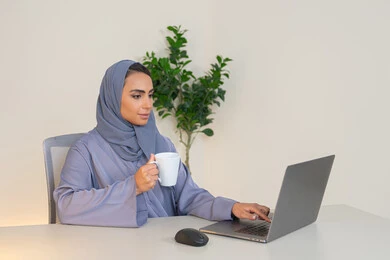 سيدة سعودية تستخدم الحاسوب في المكتب، استخدام جهاز تقني حديث ومتطور، احتساء المشروب المفضل، ايماءات التركيز والاندماج، ادارة الأعمال وانجاز المهام المطلوبة، مهنة ووظيفة نسائية،  إمرأة عربية خليجية اماراتية محجبة ترتدي عباءة ملونة تجلس خلف الطاولة وتشرب القهوة
