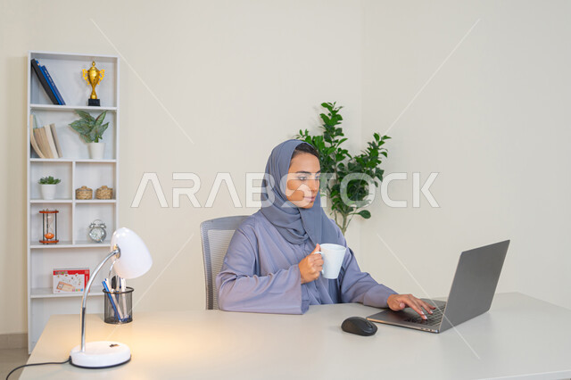 احتساء المشروب المفضل، ايماءات التركيز والاندماج، ادارة الأعمال وانجاز المهام المطلوبة، مهنة ووظيفة نسائية،  إمرأة عربية خليجية اماراتية محجبة ترتدي عباءة ملونة تجلس خلف الطاولة وتشرب القهوة، سيدة سعودية تستخدم الحاسوب في المكتب، استخدام جهاز تقني حديث ومتطور