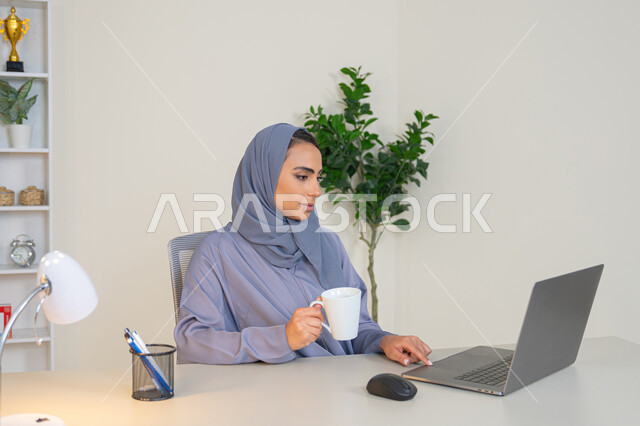احتساء المشروب المفضل، ايماءات التركيز والاندماج، ادارة الأعمال وانجاز المهام المطلوبة، مهنة ووظيفة نسائية،  إمرأة عربية خليجية اماراتية محجبة ترتدي عباءة ملونة تجلس خلف الطاولة وتشرب القهوة، سيدة سعودية تستخدم الحاسوب في المكتب، استخدام جهاز تقني حديث ومتطور