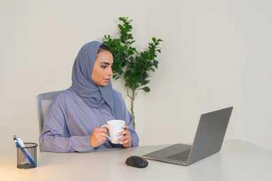 Business administration and completing required tasks, a female profession and job, a veiled Emirati Gulf Arab woman wearing a colorful abaya sitting behind a table, a Saudi woman using a computer in the office, using a modern and advanced technical device, gestures of concentration and integration