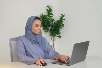 ايماءات التركيز والاندماج، ادارة الأعمال وانجاز المهام المطلوبة، مهنة ووظيفة نسائية،  إمرأة عربية خليجية اماراتية محجبة ترتدي عباءة ملونة تجلس خلف الطاولة، سيدة سعودية تستخدم الحاسوب في المكتب، استخدام جهاز تقني حديث ومتطور
