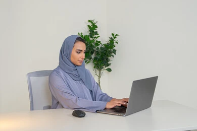 A female profession and job, a veiled Emirati Gulf Arab woman wearing a colorful abaya sitting behind a table, a Saudi woman using a computer in the office, using a modern and advanced technical device, gestures of concentration and integration, business management and completing required tasks