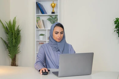 ادارة الأعمال وانجاز المهام المطلوبة، مهنة ووظيفة نسائية،  إمرأة عربية خليجية اماراتية محجبة ترتدي عباءة ملونة تجلس خلف الطاولة، سيدة سعودية تستخدم الحاسوب في المكتب، استخدام جهاز تقني حديث ومتطور، ايماءات التركيز والاندماج