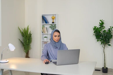 A female profession and job, a veiled Emirati Gulf Arab woman wearing a colorful abaya sitting behind a table, a Saudi woman using a computer in the office, using a modern and advanced technical device, gestures of concentration and integration, business management and completing required tasks