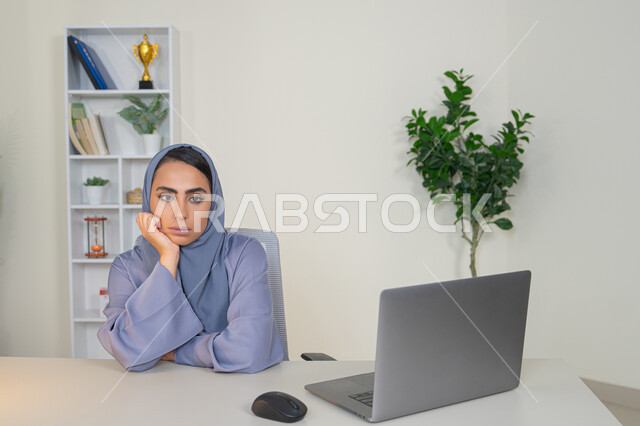 محاولة ايجاد حل لمشكلة ما, شرود الذهن والتفكير، مهنة ووظيفة مكتبية، العمل في شركات الخليج العربي، الشعور بالضجر والملل، سيدة اماراتية تستخدم التقنية الحديثة المتطورة في المكتب، امراة عربية خليجية سعودية محجبة ترتدي العباءة تجلس خلف الطاولة بإيماءات الملل