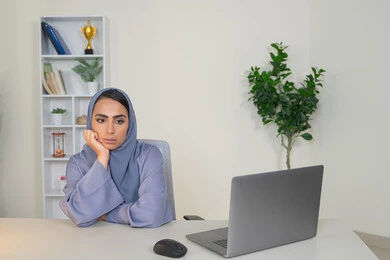محاولة ايجاد حل لمشكلة ما, شرود الذهن والتفكير، مهنة ووظيفة مكتبية، العمل في شركات الخليج العربي، الشعور بالضجر والملل، سيدة اماراتية تستخدم التقنية الحديثة المتطورة في المكتب، امراة عربية خليجية سعودية محجبة ترتدي العباءة تجلس خلف الطاولة بإيماءات الملل