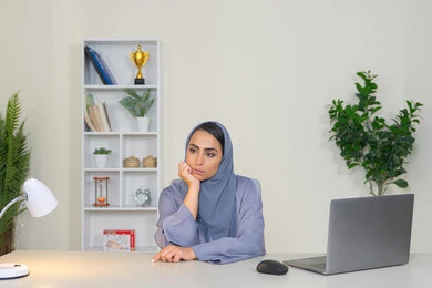 Working in a Gulf company, feeling bored, an Emirati woman using modern advanced technology in the office, a veiled Saudi Arabian Gulf woman wearing an abaya sitting behind a table with boredom, trying to find a solution to a problem, absent-mindedness and thinking, an office profession and job