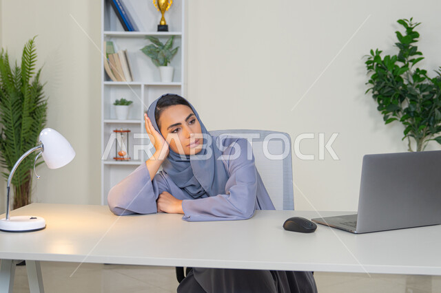 محاولة ايجاد حل لمشكلة ما, شرود الذهن والتفكير، مهنة ووظيفة مكتبية، العمل في شركات الخليج العربي، الشعور بالضجر والملل، سيدة اماراتية تستخدم التقنية الحديثة المتطورة في المكتب، امراة عربية خليجية سعودية محجبة ترتدي العباءة تجلس خلف الطاولة بإيماءات الملل