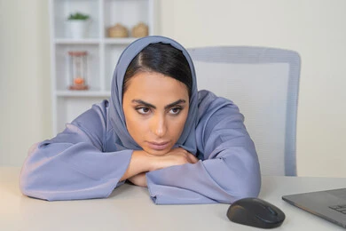 امراة عربية خليجية سعودية محجبة ترتدي العباءة تجلس خلف الطاولة بإيماءات الملل, محاولة ايجاد حل لمشكلة ما, شرود الذهن والتفكير، مهنة ووظيفة مكتبية، العمل في شركات الخليج العربي، الشعور بالضجر والملل، سيدة اماراتية تستخدم التقنية الحديثة المتطورة في المكتب