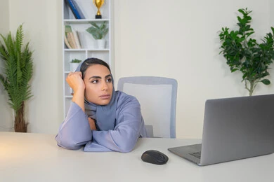 محاولة ايجاد حل لمشكلة ما, شرود الذهن والتفكير، مهنة ووظيفة مكتبية، العمل في شركات الخليج العربي، الشعور بالضجر والملل، سيدة اماراتية تستخدم التقنية الحديثة المتطورة في المكتب، امراة عربية خليجية سعودية محجبة ترتدي العباءة تجلس خلف الطاولة بإيماءات الملل