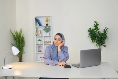 امراة عربية خليجية سعودية محجبة ترتدي العباءة تجلس خلف الطاولة بإيماءات الملل, محاولة ايجاد حل لمشكلة ما, شرود الذهن والتفكير، مهنة ووظيفة مكتبية، العمل في شركات الخليج العربي، الشعور بالضجر والملل، سيدة اماراتية تستخدم التقنية الحديثة المتطورة في المكتب
