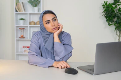 محاولة ايجاد حل لمشكلة ما, شرود الذهن والتفكير، مهنة ووظيفة مكتبية، العمل في شركات الخليج العربي، الشعور بالضجر والملل، سيدة اماراتية تستخدم التقنية الحديثة المتطورة في المكتب، امراة عربية خليجية سعودية محجبة ترتدي العباءة تجلس خلف الطاولة بإيماءات الملل