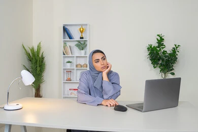 سيدة اماراتية تستخدم التقنية الحديثة المتطورة في المكتب، امراة عربية خليجية سعودية محجبة ترتدي العباءة تجلس خلف الطاولة بإيماءات الملل, محاولة ايجاد حل لمشكلة ما, شرود الذهن والتفكير، مهنة ووظيفة مكتبية، العمل في شركات الخليج العربي، الشعور بالضجر والملل