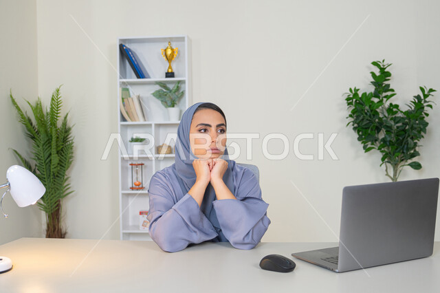 امراة عربية خليجية سعودية محجبة ترتدي العباءة تجلس خلف الطاولة بإيماءات الملل, محاولة ايجاد حل لمشكلة ما, شرود الذهن والتفكير، مهنة ووظيفة مكتبية، العمل في شركات الخليج العربي، الشعور بالضجر والملل، سيدة اماراتية تستخدم التقنية الحديثة المتطورة في المكتب