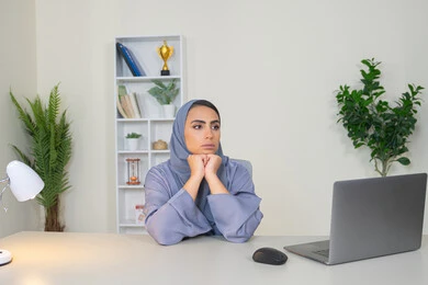 A veiled Saudi Arabian Gulf woman wearing an abaya sitting behind a table with bored gestures, trying to find a solution to a problem, absent-minded and thinking, an office job and profession, working in Gulf companies, feeling bored and fed up, an Emirati woman using modern advanced technology in the office
