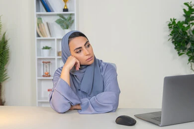 سيدة اماراتية تستخدم التقنية الحديثة المتطورة في المكتب، امراة عربية خليجية سعودية محجبة ترتدي العباءة تجلس خلف الطاولة بإيماءات الملل, محاولة ايجاد حل لمشكلة ما, شرود الذهن والتفكير، مهنة ووظيفة مكتبية، العمل في شركات الخليج العربي، الشعور بالضجر والملل