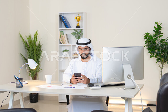 إدارة شؤون الأعمال وتنظيمها عبر الجوال، رجل عربي خليجي اماراتي يرتدي الكندورة والغترة البيضاء يجلس خلف الطاولة ويمسك بيده الهاتف المحمول، استخدام جهاز تقني حديث ومتطور، انجاز مهام العمل في المكتب، تصفح التطبيقات وبرامج التواصل الاجتماعي، وظيفة ومهنة مكتبية