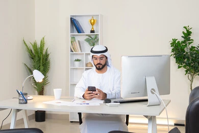 Managing and organizing business affairs via mobile, an Emirati Gulf Arab man wearing a white kandura and ghutra sitting behind a table holding a mobile phone, using a modern and advanced technical device, completing work tasks in the office, browsing applications and social networking programs, an office job and profession