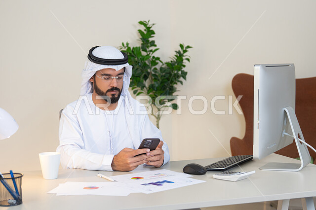 إدارة شؤون الأعمال وتنظيمها عبر الجوال، رجل عربي خليجي اماراتي يرتدي الكندورة والغترة البيضاء يجلس خلف الطاولة ويمسك بيده الهاتف المحمول، استخدام جهاز تقني حديث ومتطور، انجاز مهام العمل في المكتب، تصفح التطبيقات وبرامج التواصل الاجتماعي، وظيفة ومهنة مكتبية