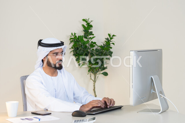 دمج العمل مع التقنية والتكنولوجيا، تطور الشركات الاماراتية، إتمام مهام العمل في المكتب، إدارة شؤون الأعمال وتنظيمها، احتساء مشروب القهوة، استخدام جهاز تقني حديث ومتطور، رجل عربي خليجي اماراتي يرتدي الكندورة والغترة البيضاء يستخدم الحاسوب، مهنة ووظيفة مكتبية
