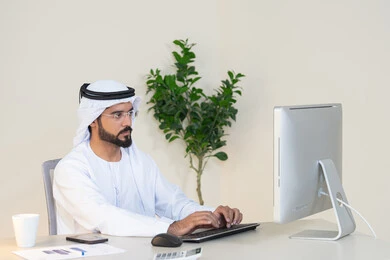 دمج العمل مع التقنية والتكنولوجيا، تطور الشركات الاماراتية، إتمام مهام العمل في المكتب، إدارة شؤون الأعمال وتنظيمها، احتساء مشروب القهوة، استخدام جهاز تقني حديث ومتطور، رجل عربي خليجي اماراتي يرتدي الكندورة والغترة البيضاء يستخدم الحاسوب، مهنة ووظيفة مكتبية