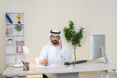 Sipping your favorite drink at the office, an Emirati Gulf Arab man wearing a white kandura and ghutra sitting behind a table holding a mobile phone and a cup of coffee next to him, working in a UAE company, an office job and profession, making a phone call, using a modern and advanced technical device