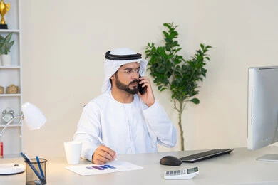 اجراء مكالمة هاتفية، استخدام جهاز تقني حديث ومتطور، احتساء المشروب المفضل في المكتب، رجل عربي خليجي اماراتي يرتدي الكندورة والغترة البيضاء يجلس خلف الطاولة يمسك بيده الهاتف المحمول وبجانبه كوب من القهوة، العمل في شركات الامارات العربية المتحدة، مهنة ووظيفة مكتبية