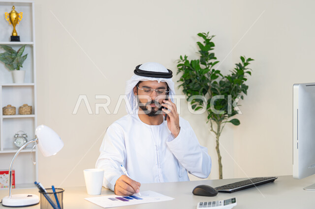 العمل في شركات الامارات العربية المتحدة، مهنة ووظيفة مكتبية، اجراء مكالمة هاتفية، استخدام جهاز تقني حديث ومتطور، احتساء المشروب المفضل في المكتب، رجل عربي خليجي اماراتي يرتدي الكندورة والغترة البيضاء يجلس خلف الطاولة يمسك بيده الهاتف المحمول وبجانبه كوب من القهوة