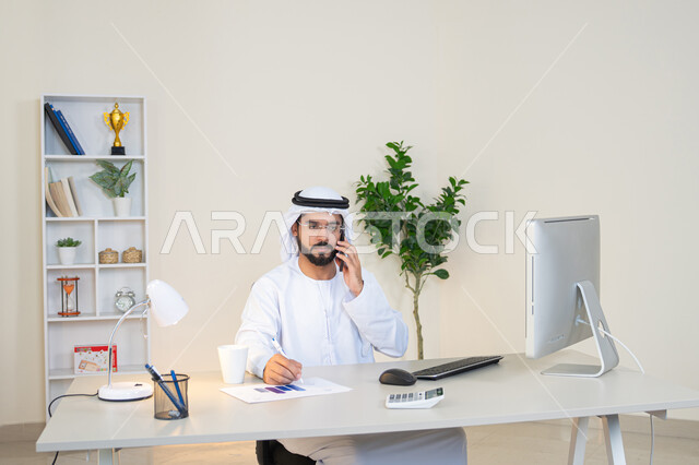 احتساء المشروب المفضل في المكتب، رجل عربي خليجي اماراتي يرتدي الكندورة والغترة البيضاء يجلس خلف الطاولة يمسك بيده الهاتف المحمول وبجانبه كوب من القهوة، العمل في شركات الامارات العربية المتحدة، مهنة ووظيفة مكتبية، اجراء مكالمة هاتفية، استخدام جهاز تقني حديث ومتطور