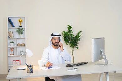 احتساء المشروب المفضل في المكتب، رجل عربي خليجي اماراتي يرتدي الكندورة والغترة البيضاء يجلس خلف الطاولة يمسك بيده الهاتف المحمول وبجانبه كوب من القهوة، العمل في شركات الامارات العربية المتحدة، مهنة ووظيفة مكتبية، اجراء مكالمة هاتفية، استخدام جهاز تقني حديث ومتطور