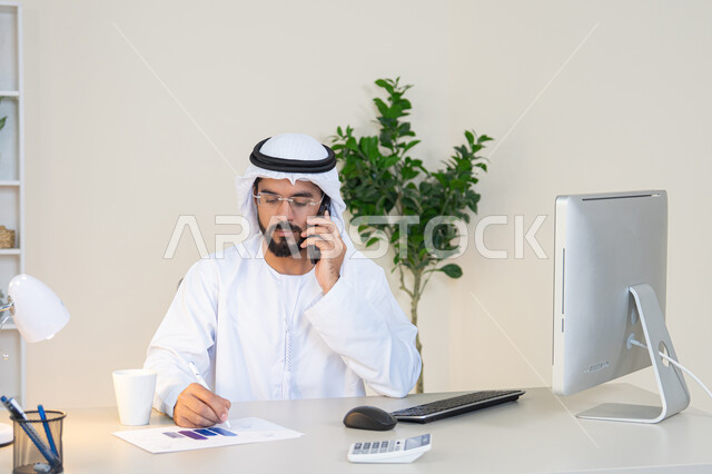 اجراء مكالمة هاتفية، استخدام جهاز تقني حديث ومتطور، احتساء المشروب المفضل في المكتب، رجل عربي خليجي اماراتي يرتدي الكندورة والغترة البيضاء يجلس خلف الطاولة يمسك بيده الهاتف المحمول وبجانبه كوب من القهوة، العمل في شركات الامارات العربية المتحدة، مهنة ووظيفة مكتبية