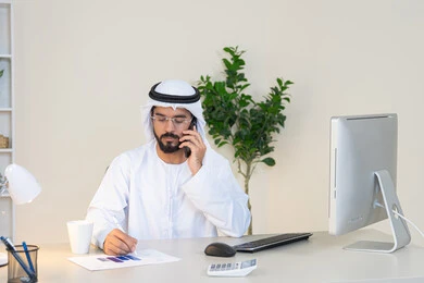 اجراء مكالمة هاتفية، استخدام جهاز تقني حديث ومتطور، احتساء المشروب المفضل في المكتب، رجل عربي خليجي اماراتي يرتدي الكندورة والغترة البيضاء يجلس خلف الطاولة يمسك بيده الهاتف المحمول وبجانبه كوب من القهوة، العمل في شركات الامارات العربية المتحدة، مهنة ووظيفة مكتبية