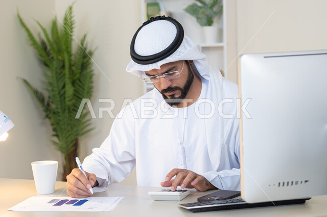 تحليل نتائج البيانات الاحصائية، رجل عربي خليجي اماراتي يرتدي الكندورة والغترة يجلس خلف الطاولة في المكتب ويعمل على الحاسب المحمول لانجاز المهام، تدوين الملاحظات على الورق، استخدام الالة الحاسبة لاجراء العمليات الحسابية، وظيفة مكتبية في شركات الامارات العربية المتحدة