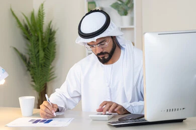 تحليل نتائج البيانات الاحصائية، رجل عربي خليجي اماراتي يرتدي الكندورة والغترة يجلس خلف الطاولة في المكتب ويعمل على الحاسب المحمول لانجاز المهام، تدوين الملاحظات على الورق، استخدام الالة الحاسبة لاجراء العمليات الحسابية، وظيفة مكتبية في شركات الامارات العربية المتحدة