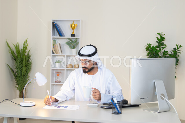 استخدام الالة الحاسبة لاجراء العمليات الحسابية، وظيفة مكتبية في شركات الامارات العربية المتحدة، تحليل نتائج البيانات الاحصائية، رجل عربي خليجي اماراتي يرتدي الكندورة والغترة يجلس خلف الطاولة في المكتب ويعمل على الحاسب المحمول لانجاز المهام، تدوين الملاحظات على الورق