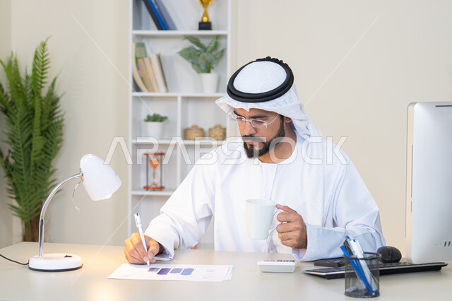تحليل نتائج البيانات الاحصائية، رجل عربي خليجي اماراتي يرتدي الكندورة والغترة يجلس خلف الطاولة في المكتب ويعمل على الحاسب المحمول لانجاز المهام، تدوين الملاحظات على الورق، استخدام الالة الحاسبة لاجراء العمليات الحسابية، وظيفة مكتبية في شركات الامارات العربية المتحدة
