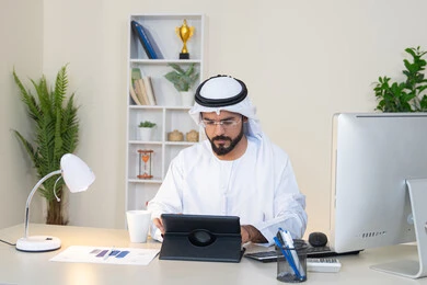 إنجاز مهام العمل، إدارة شؤون الأعمال وتنظيمها عبر الايباد، استخدام جهاز تقني حديث ومتطور، رجل عربي خليجي اماراتي يرتدي الكندورة والغترة البيضاء يجلس خلف الطاولة في المكتب ويستخدم الجهاز اللوحي، مهنة ووظيفة مكتبية