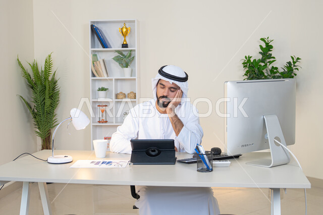 مهنة ووظيفة مكتبية، إنجاز مهام العمل، إدارة شؤون الأعمال وتنظيمها عبر الايباد، استخدام جهاز تقني حديث ومتطور، رجل عربي خليجي اماراتي يرتدي الكندورة والغترة البيضاء يضع يده على خده بايماءات الملل يجلس خلف الطاولة في المكتب ويستخدم الجهاز اللوحي