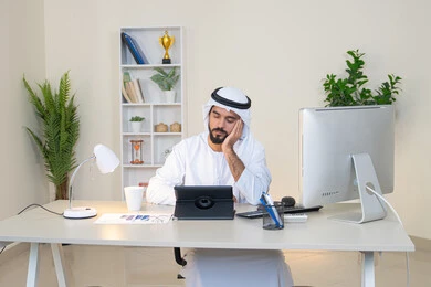 مهنة ووظيفة مكتبية، إنجاز مهام العمل، إدارة شؤون الأعمال وتنظيمها عبر الايباد، استخدام جهاز تقني حديث ومتطور، رجل عربي خليجي اماراتي يرتدي الكندورة والغترة البيضاء يضع يده على خده بايماءات الملل يجلس خلف الطاولة في المكتب ويستخدم الجهاز اللوحي