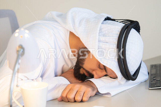 Gestures of drowsiness, fatigue and exhaustion, an Arab Gulf Emirati man wearing a white kandura and ghutra sitting behind a desk in the office taking a break, working in UAE companies, office profession and job, sleeping on the work desk